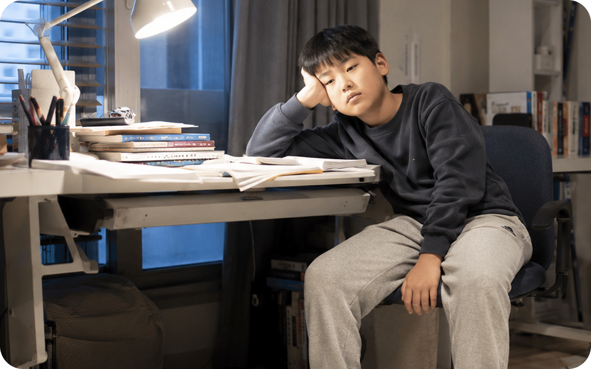 책이 쌓인 책상 앞에서 턱을 괴고 지친 표정으로 앉아 있는 아이