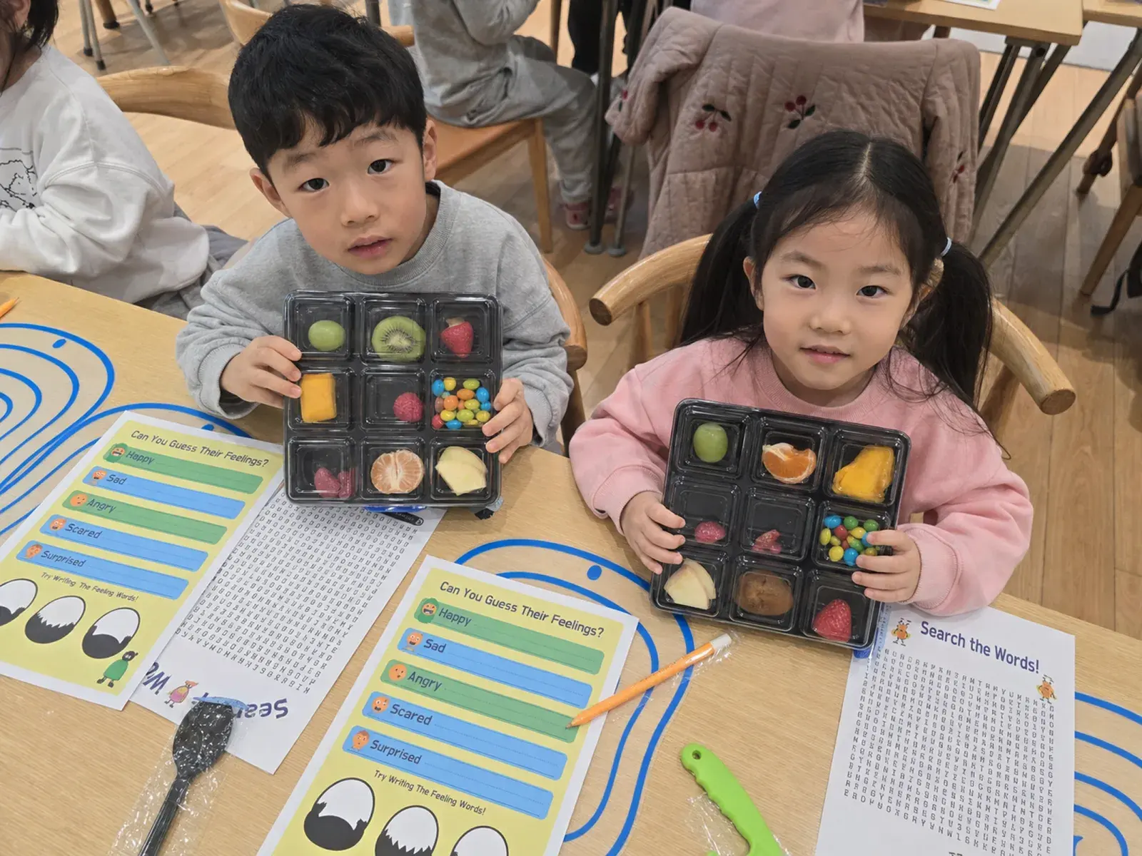 두 아이가 아홉 칸에 과일과 재료를 나눠 담은 트레이를 들어 보이고, 앞에는 영어 워크시트가 놓여 있다.
