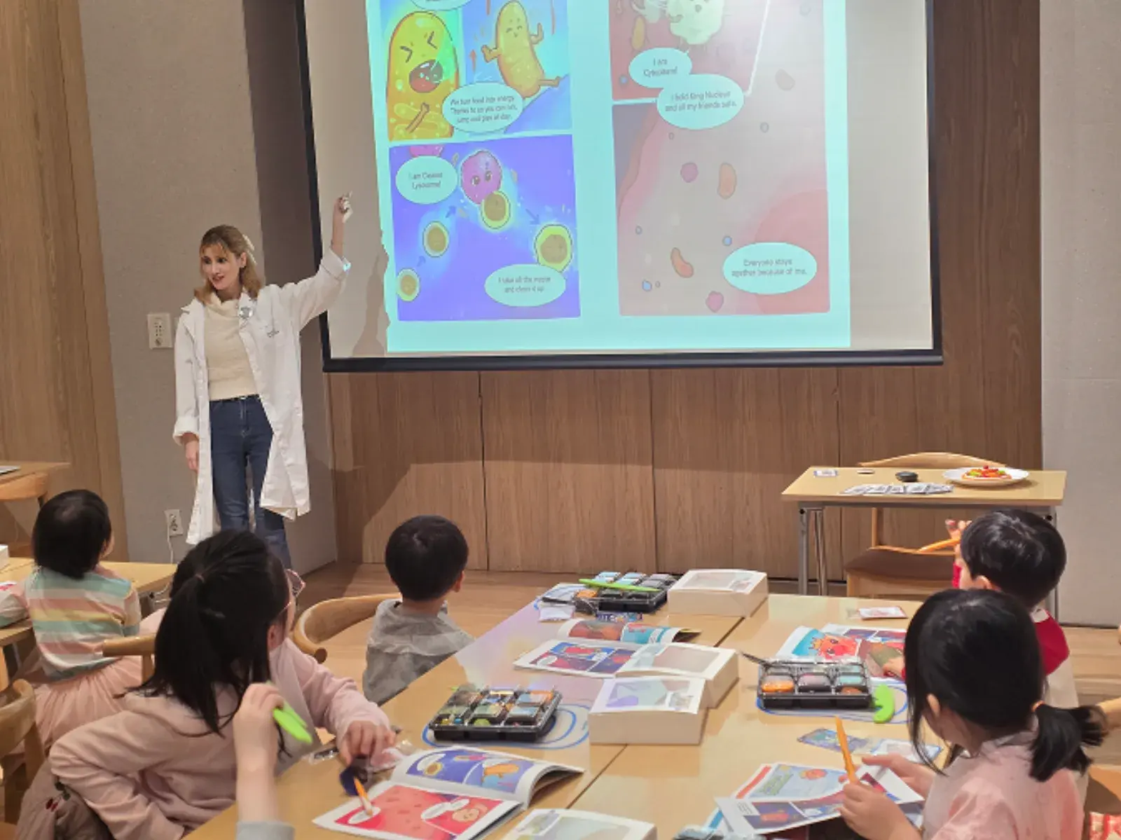 흰 가운을 입은 강사가 세포 캐릭터 만화가 띄워진 화면을 가리키고, 아이들이 각자 워크북을 펴고 보고 있다.